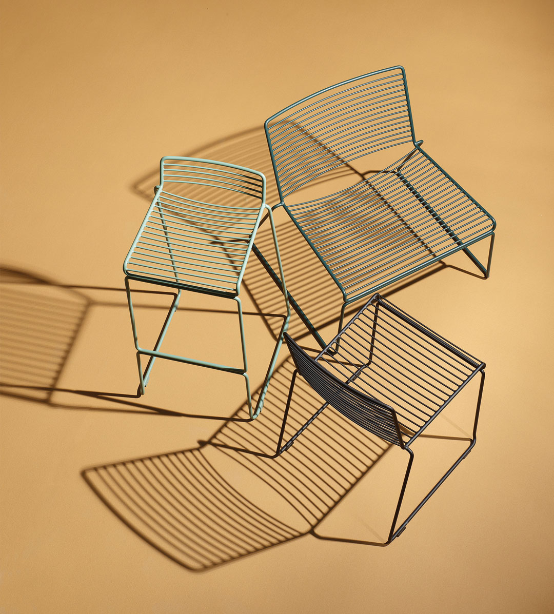 Three minimalist wireframe chairs in different heights and colours are arranged on a beige surface, casting distinct shadows—perfect inspiration for your next gathering with Trinidad Event Furniture. - Perched Event Rentals, Trinidad