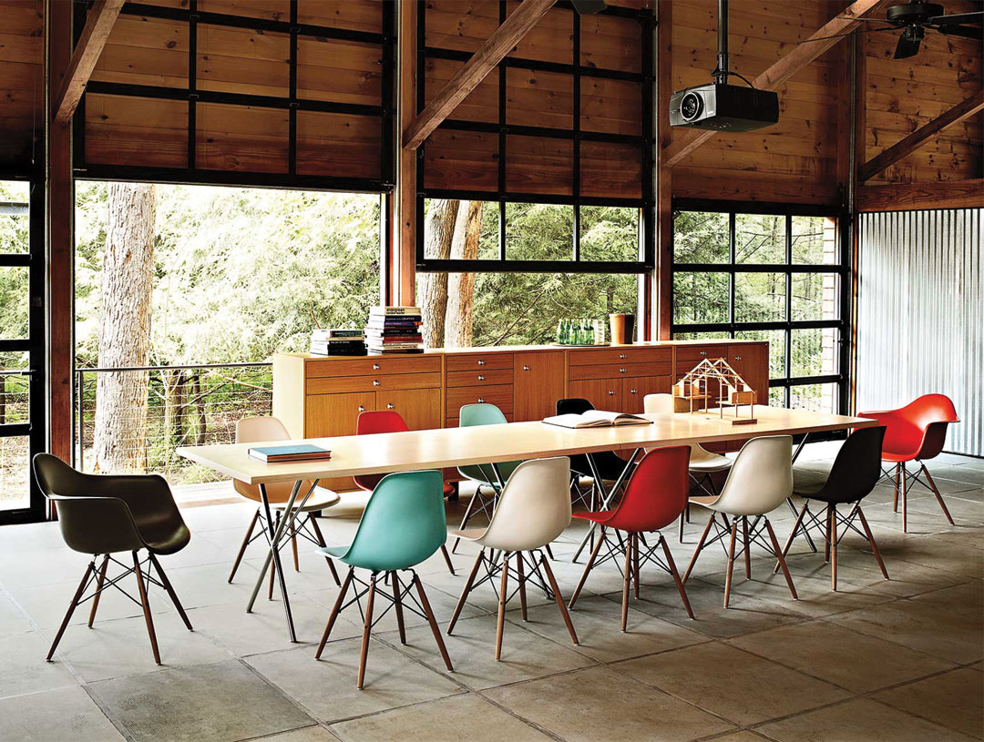 A modern conference room with a long wooden table, assorted coloured chairs from Trinidad Event Furniture, a sideboard with stacked books, and large windows showing trees outside. - Perched Event Rentals, Trinidad