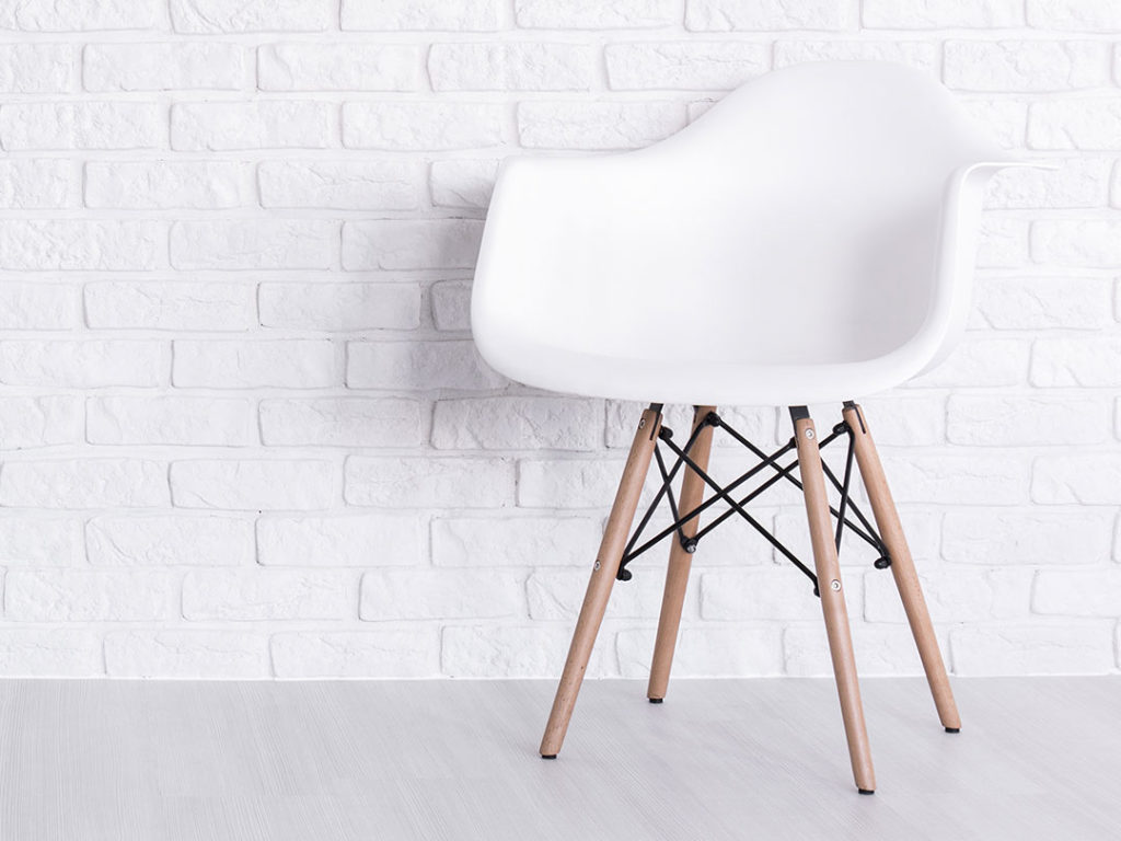 Eames Armchair, a modern white plastic chair with wooden legs, against a white brick wall on a light-coloured floor