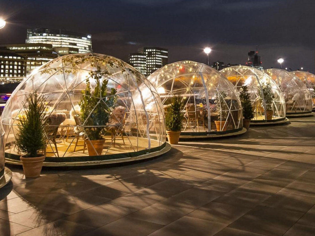 A row of Geodesic Air Conditioning Domes with tables and chairs on an outdoor terrace at night, with potted plants