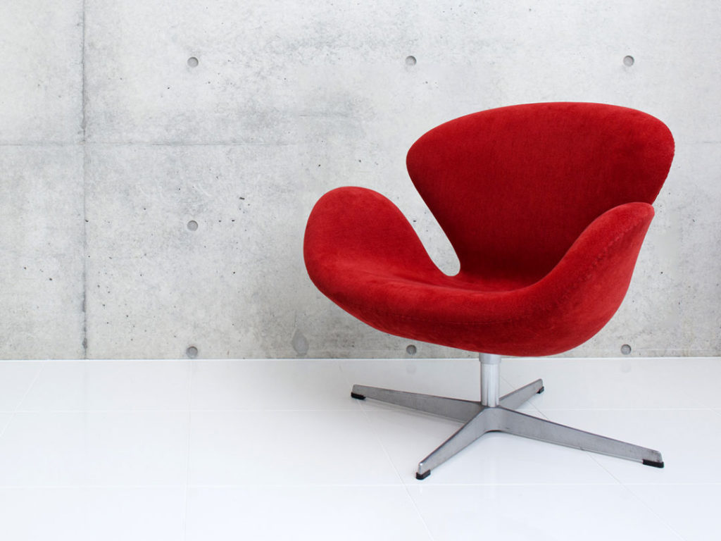 Swan Chair with a red upholstered seat, curved design and metal base, on a white floor against a grey concrete wall
