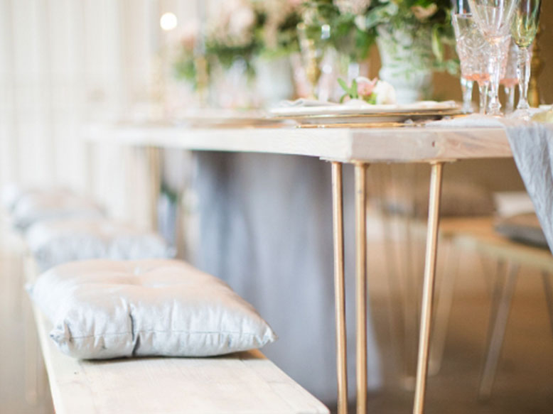 The Hairpin Table from Trinidad Event Rentals is set for dining, paired with a bench with silver seat cushions and adorned with elegant glassware and floral arrangements. - Perched Event Rentals, Trinidad