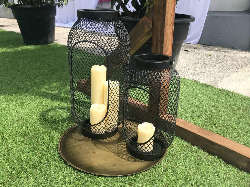 Three cream-coloured Candle Lanterns rest on a round brass tray on artificial grass
