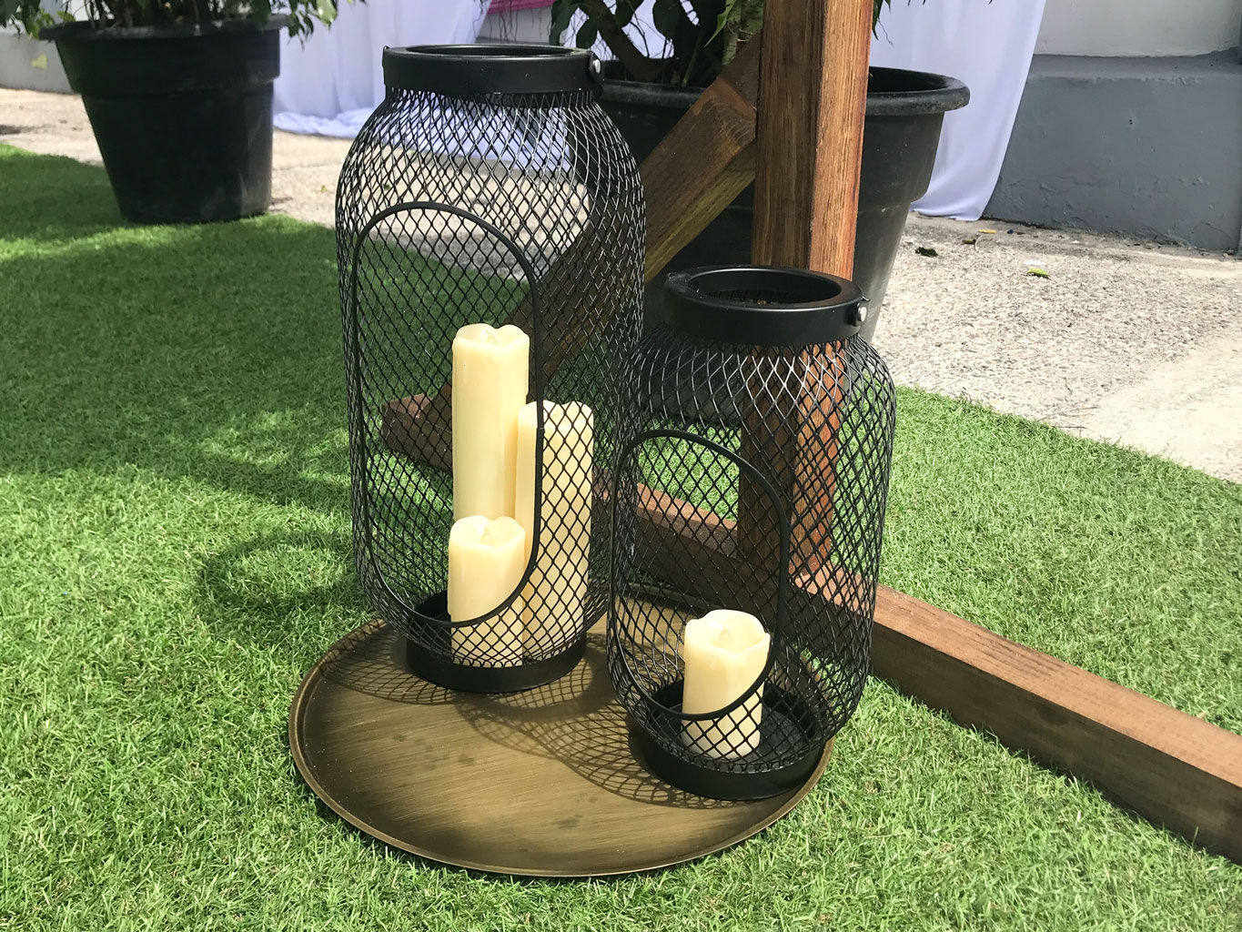 Three cream-coloured Candle Lanterns rest on a round brass tray on artificial grass