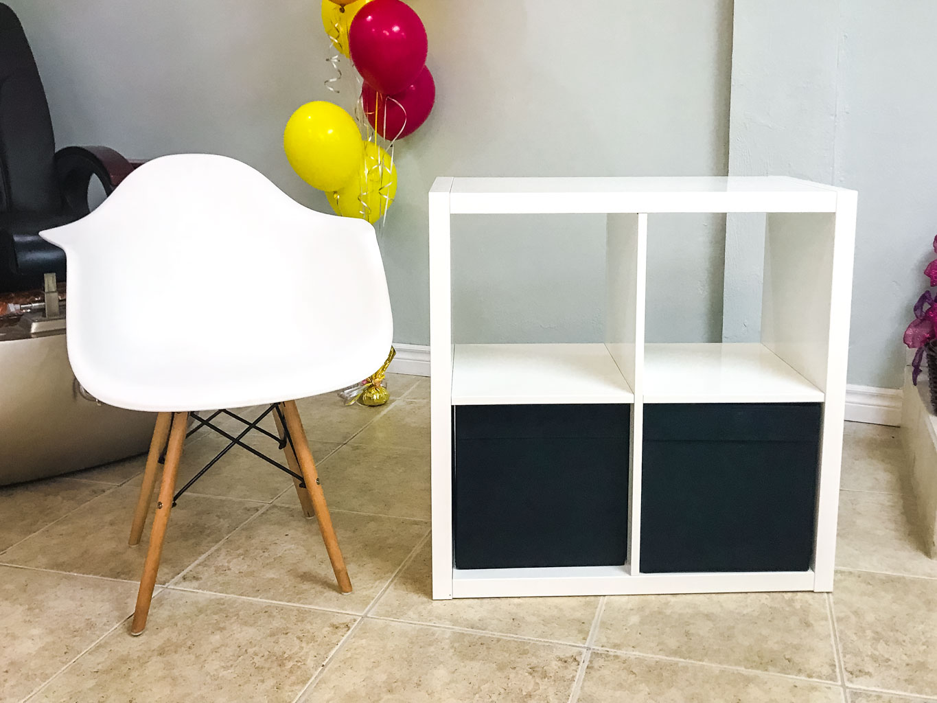 Simple Storage Table with two black bins beside a white chair with wooden legs, with red and yellow balloons behind