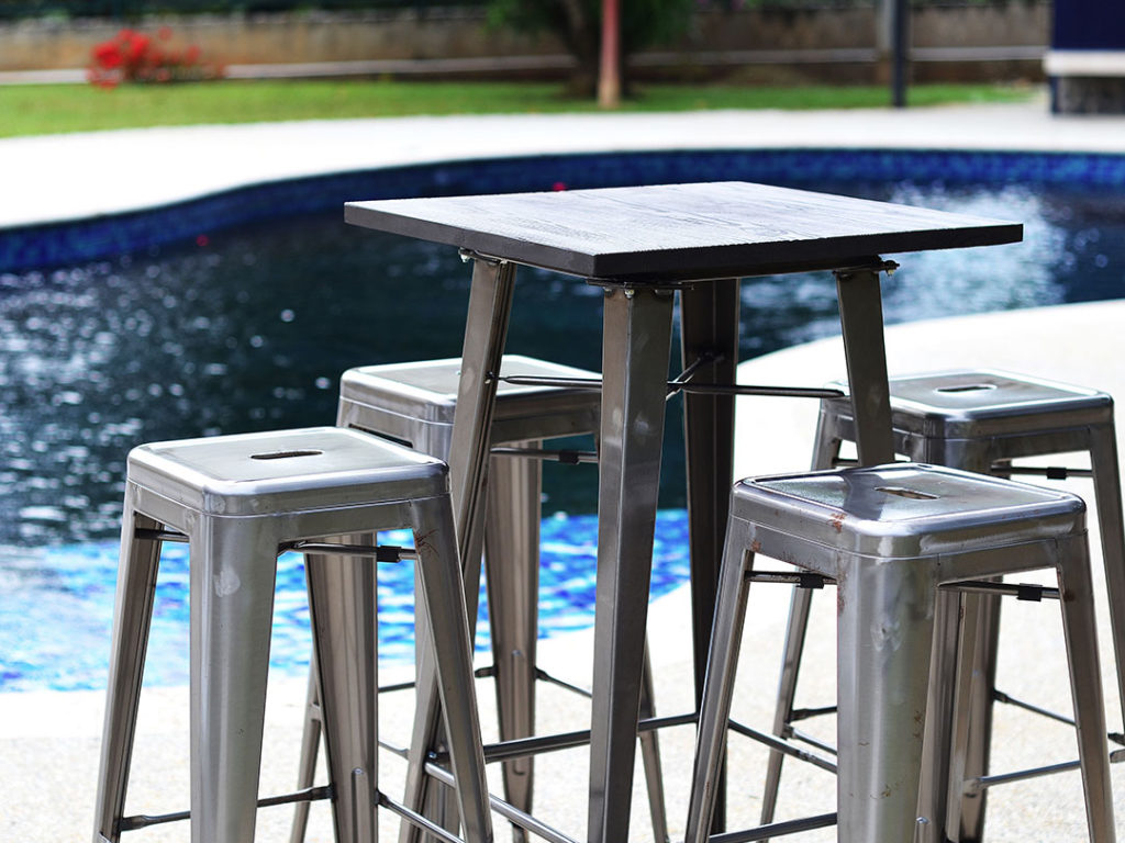 An Industrial Cocktail Table with four matching bar stools beside a swimming pool, with water visible behind