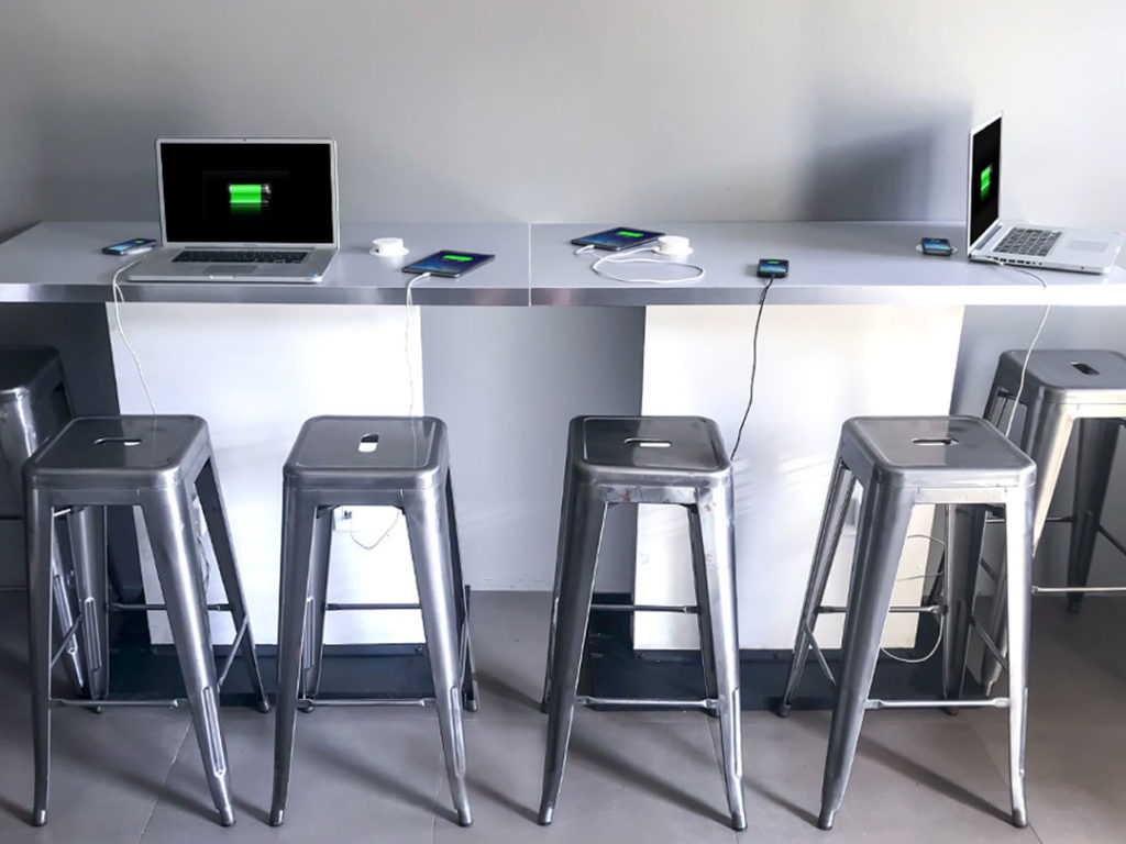 Four Wireless Charging Breakout Tables (4ft) with metal stools, each charging laptops and phones that show charging screens