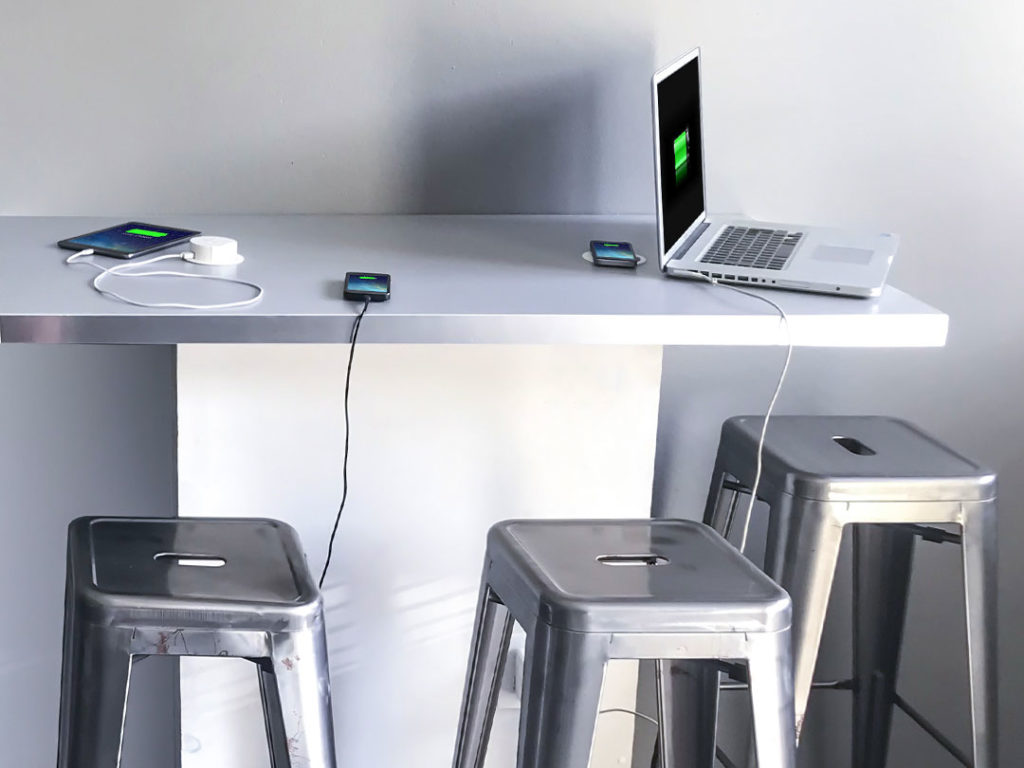 A laptop, phone and tablet charging on the Wireless Charging Breakout Table, with metal stools underneath, in a modern space