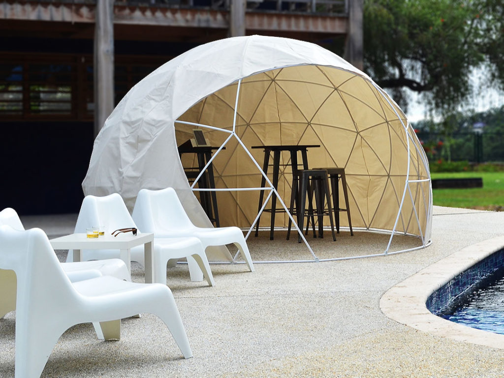Geodesic Dome Tent with stools and high tables inside, set by a pool, with white outdoor chairs arranged nearby
