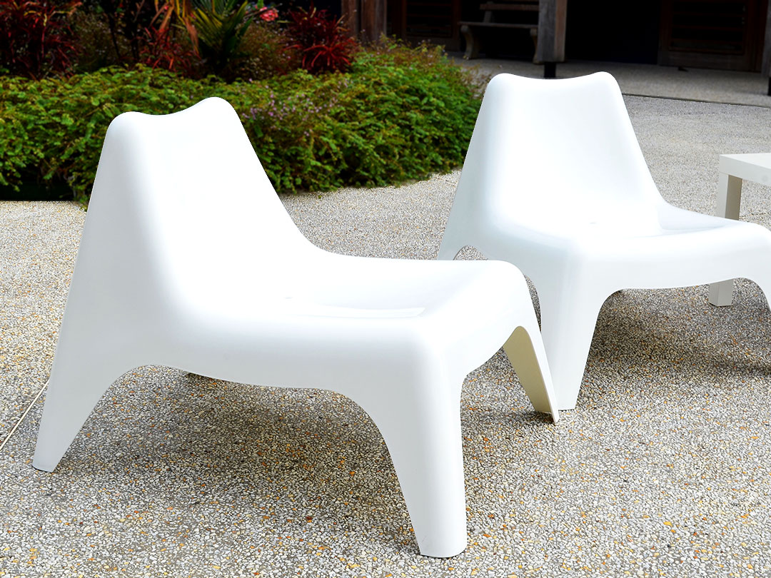 Two Poolside Easy Chairs, modern and white, on a gravel patio surrounded by green plants, with a building behind