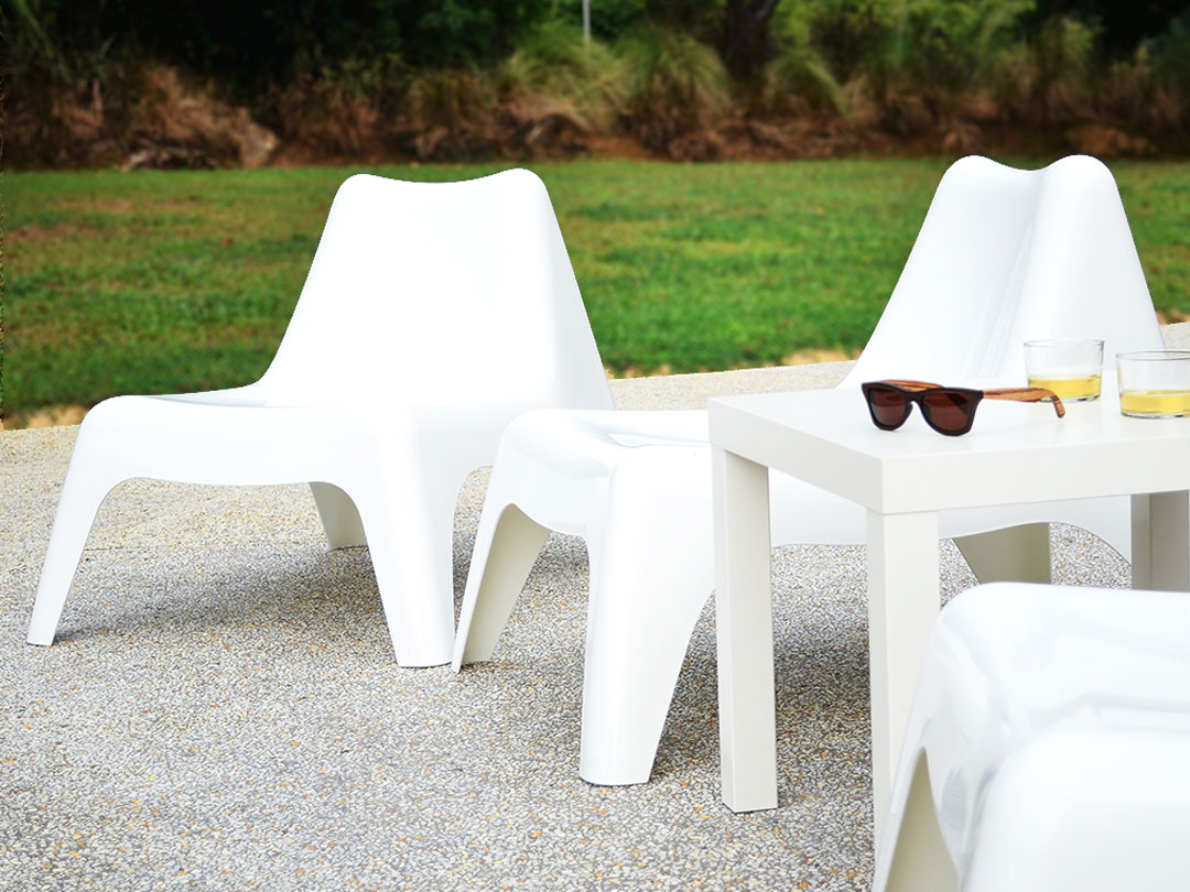 Two modern white Poolside Easy Chairs sit on a textured patio next to a sleek table with sunglasses and drinks, framed by grass and shrubs in the background. - Perched Event Rentals, Trinidad
