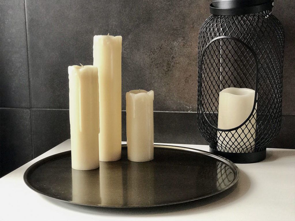 Three Flameless Candles stand on a black tray, with a fourth inside a wire lantern, set against dark tiles