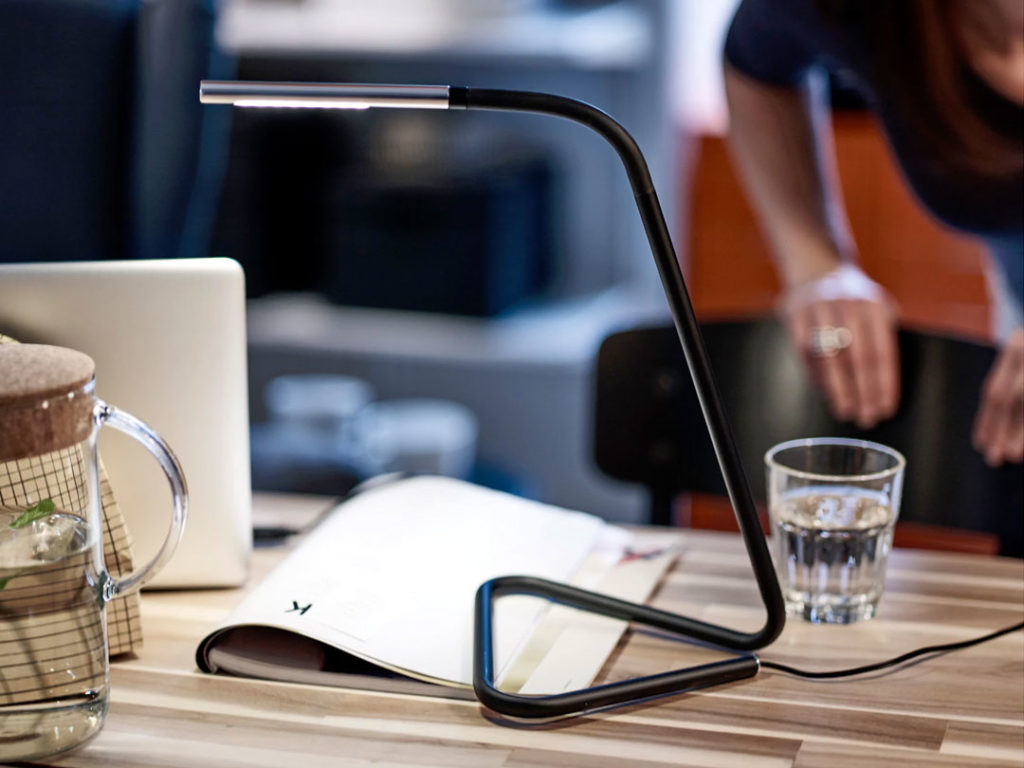 Flex Lamp, a black LED desk lamp with an adjustable head, on a wooden table beside an open magazine and a glass of water