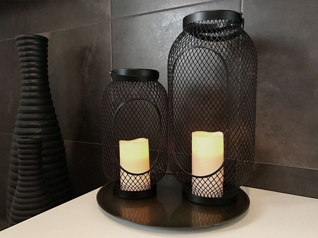 A Candle Lantern with battery-operated candles sits on a round tray next to a textured black vase, all displayed on a white surface against dark tiled walls—a stylish accent from Trinidad Wedding Hire. - Perched Event Rentals, Trinidad