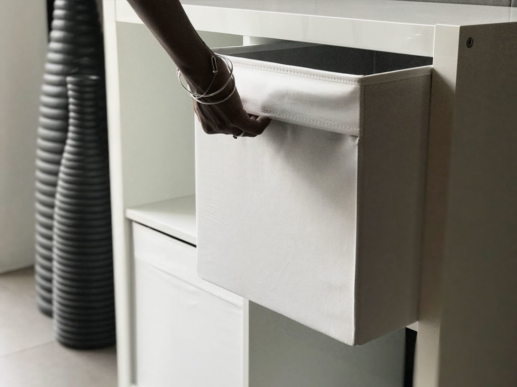 A hand pulls a Simple Storage Cube out of a white shelving unit, with two tall ribbed black vases in the background