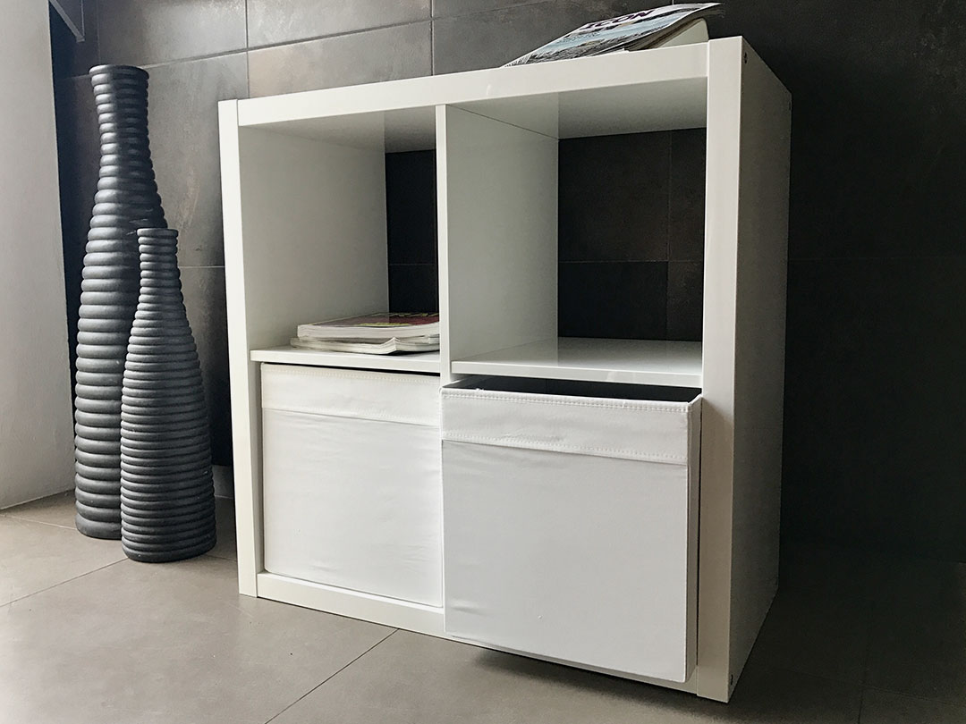 A Simple Storage Table from Trinidad Rental Furniture, featuring two fabric bins and a stack of magazines on top, sits on a tiled floor beside two tall, striped vases. - Perched Event Rentals, Trinidad