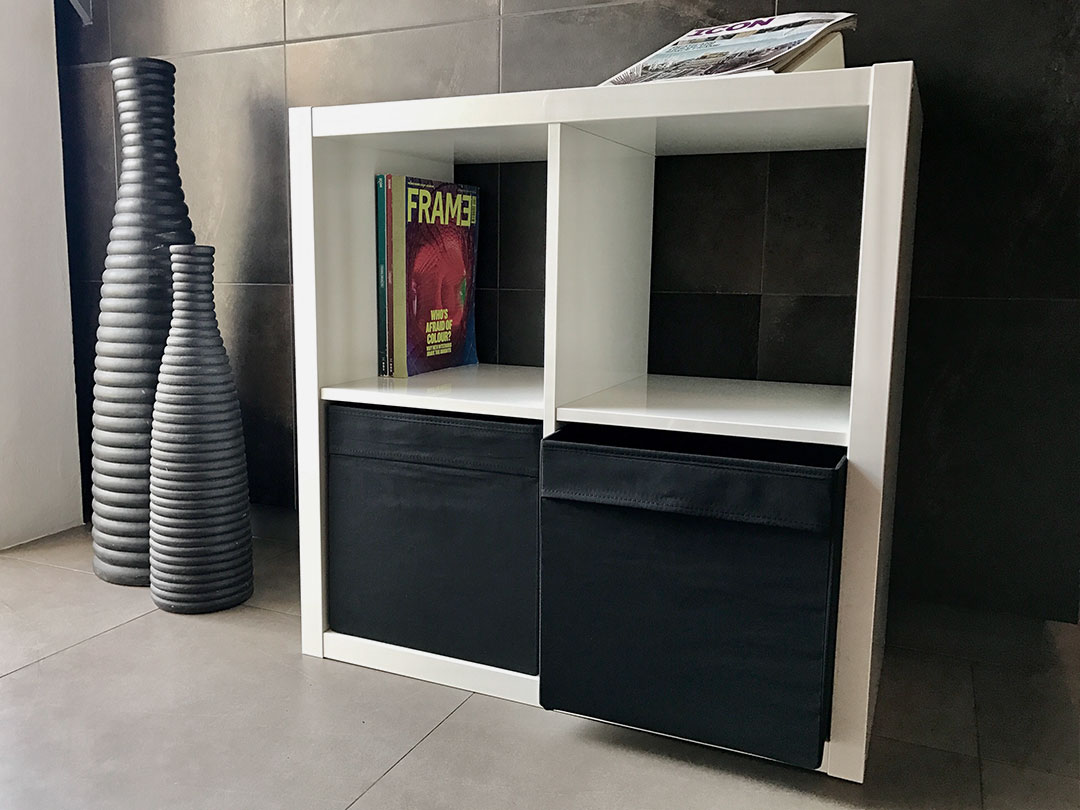The Simple Storage Table, a white cubical shelf with magazines and black fabric bins, stands on a tiled floor beside ribbed vases and a tiled wall—an ideal choice for stylish Trinidad Rental Furniture. - Perched Event Rentals, Trinidad