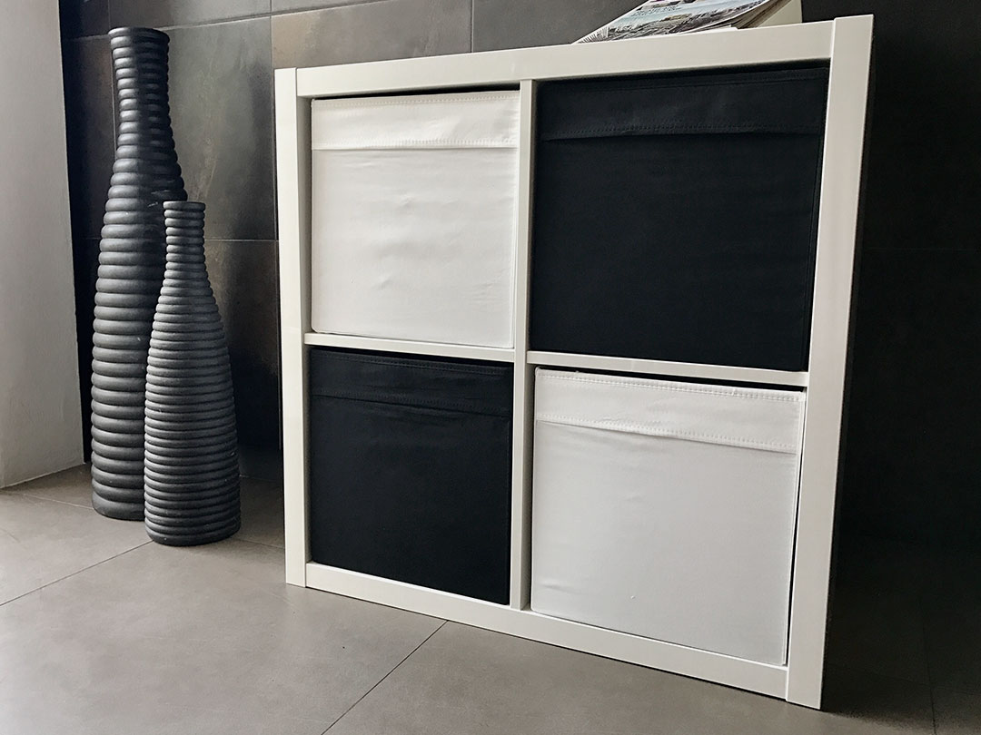 The Simple Storage Table, a white cube unit with four fabric bins—two black, two white—stands on tiles beside three tall black ribbed vases, ideal for modern décor from Trinidad Event Rentals. - Perched Event Rentals, Trinidad