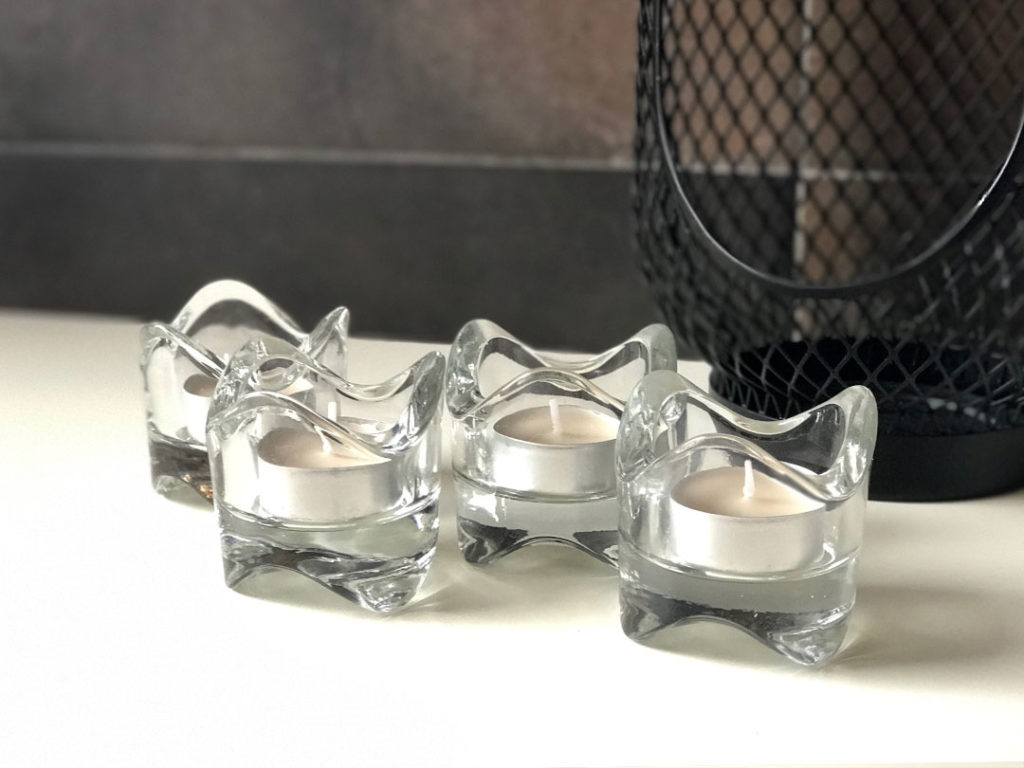 Four Wave Tealight glass candle holders with white candles, arranged on a white surface beside a black mesh décor piece