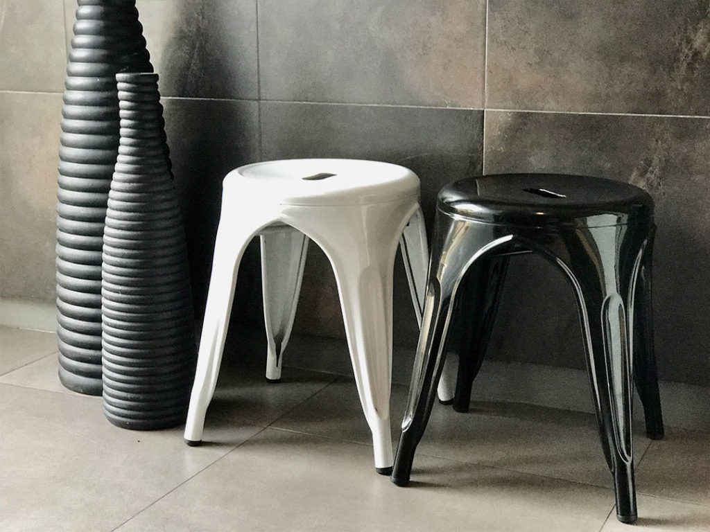 Two Industrial Stools, one white and one black, side by side next to three tall, ribbed vases against a tiled wall