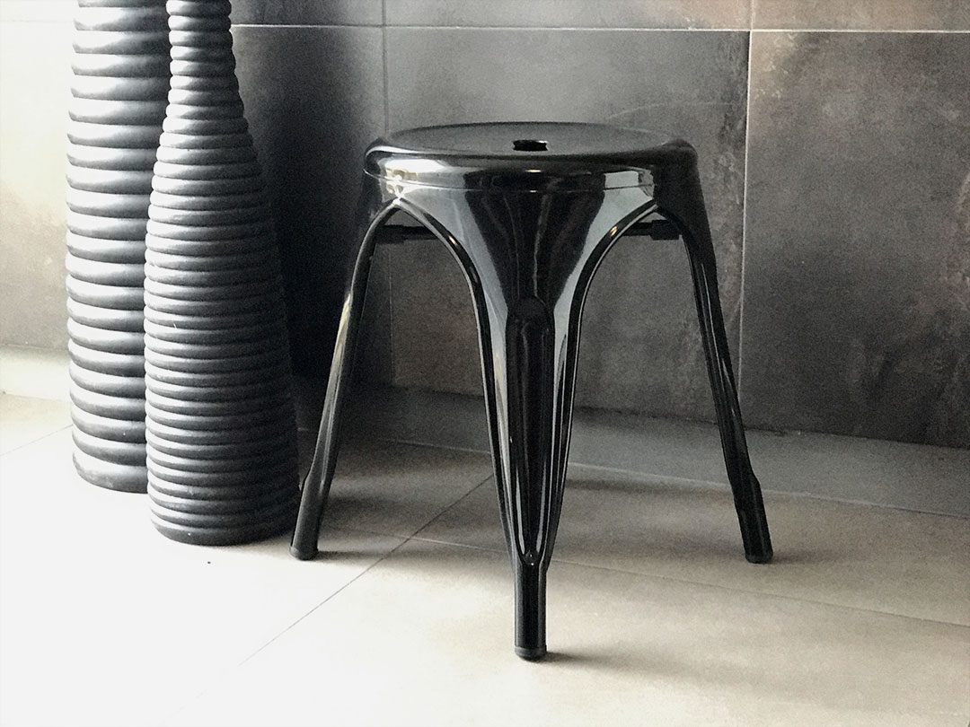 The Industrial Stool, a glossy black three-legged seat, sits on tiled flooring beside tall ribbed black vases against a dark tiled wall—ideal for stylish interiors from Trinidad Event Hire. - Perched Event Rentals, Trinidad