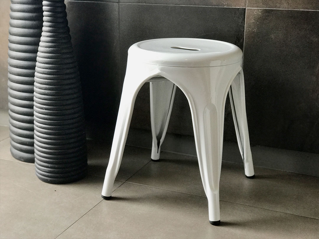 An industrial stool in white metal with four legs is placed on a tiled floor beside two tall, textured black vases—perfect for elevating elegant spaces with Trinidad Event Rentals. - Perched Event Rentals, Trinidad