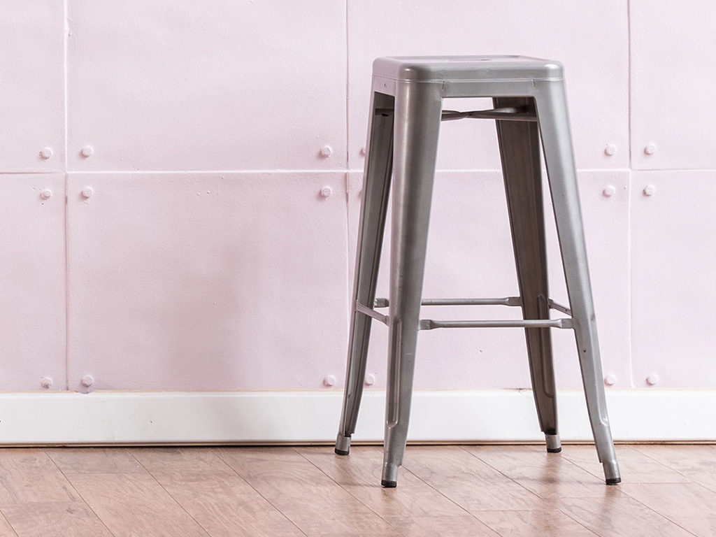 An Industrial Bar Stool sits on a wooden floor in front of a light pink, riveted wall
