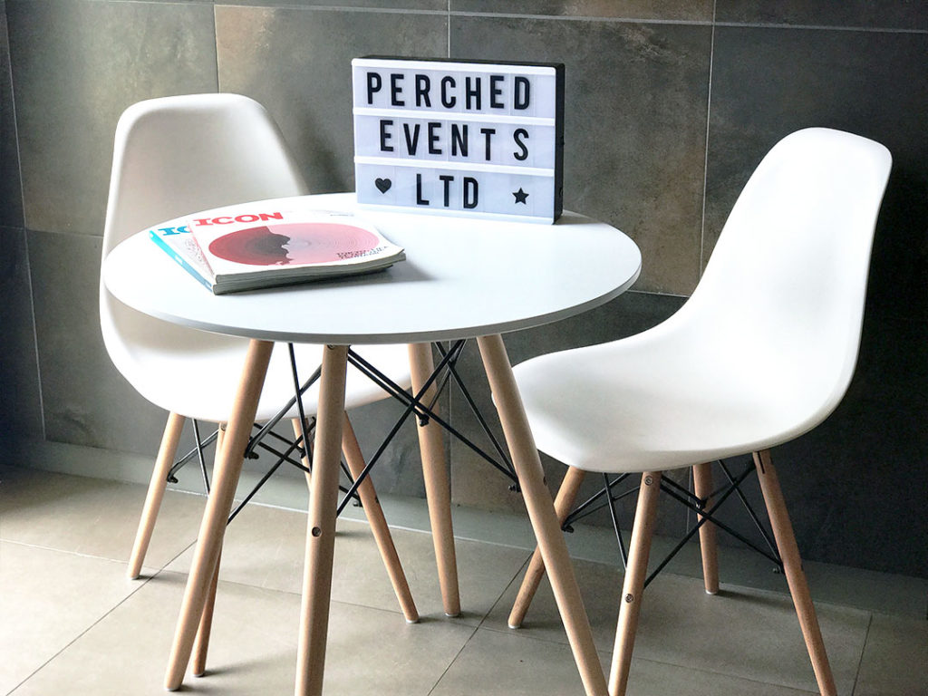 Eames Table, a small round table with two white chairs, magazines on the table and a sign reading PERCHED EVENTS LTD behind