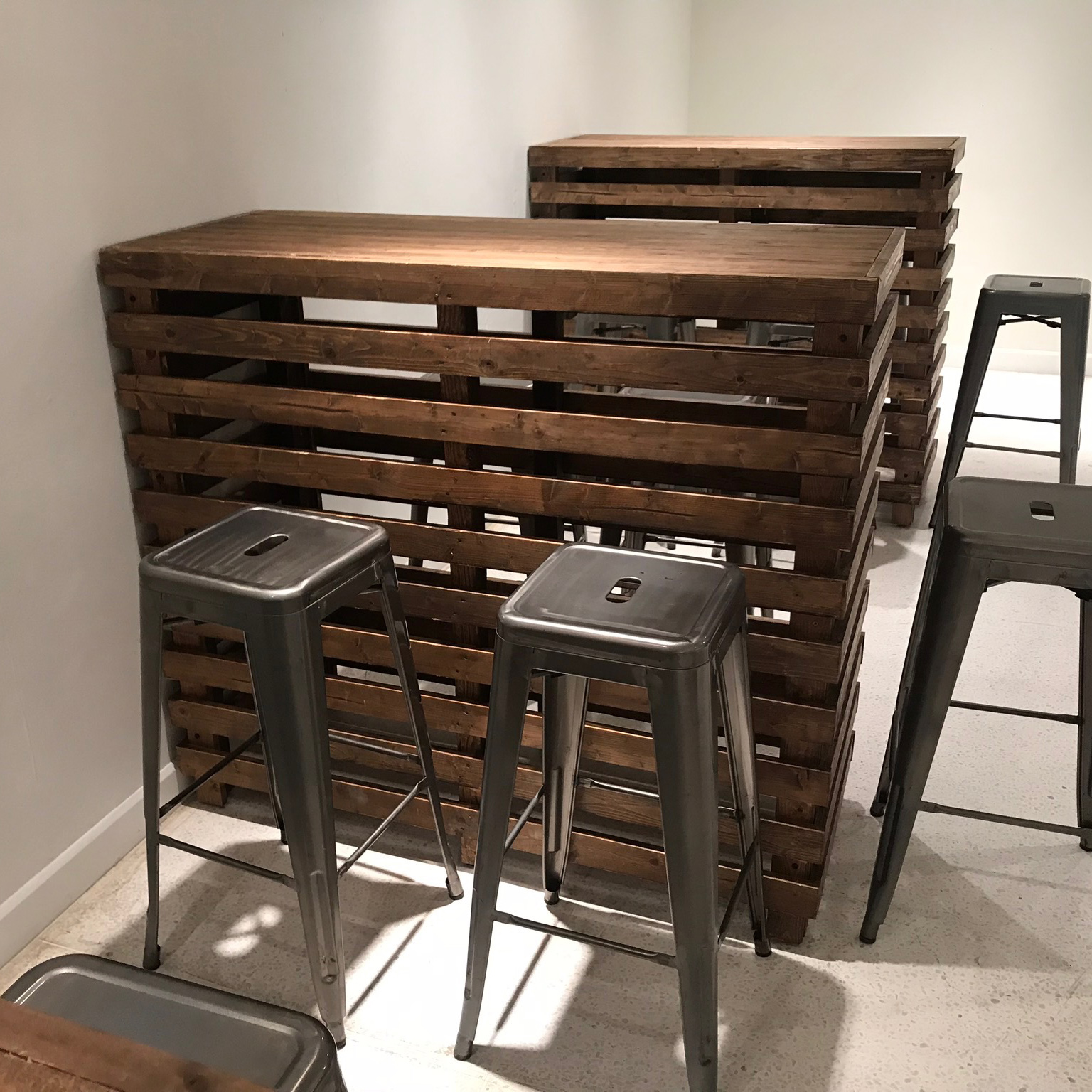 Pallet Bar Tables paired with metal stools are set on a light floor in a minimalist space—ideal for Trinidad Event Hire and stylish gatherings. - Perched Event Rentals, Trinidad