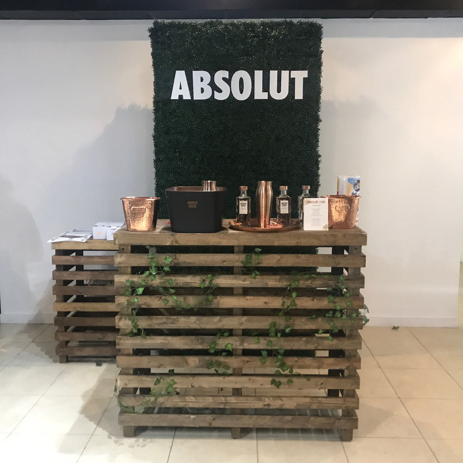 A Pallet Bar Table with copper drinkware and Absolut vodka bottles stands before a green faux hedge wall featuring the "ABSOLUT" logo, presenting stylish event options from Trinidad Event Furniture. - Perched Event Rentals, Trinidad