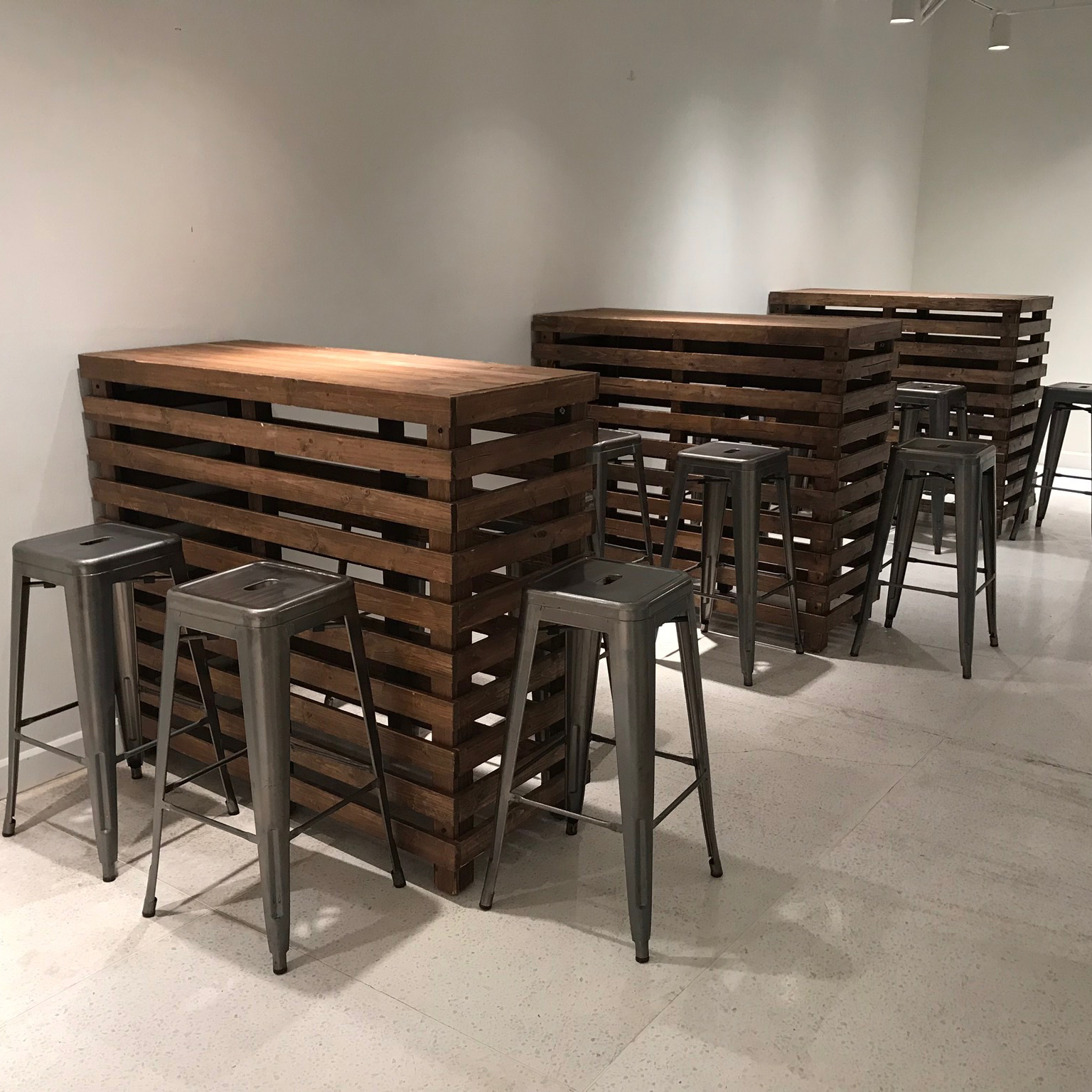 Four Pallet Bar Tables, each paired with metal stools, arranged in a room with light-coloured walls and flooring