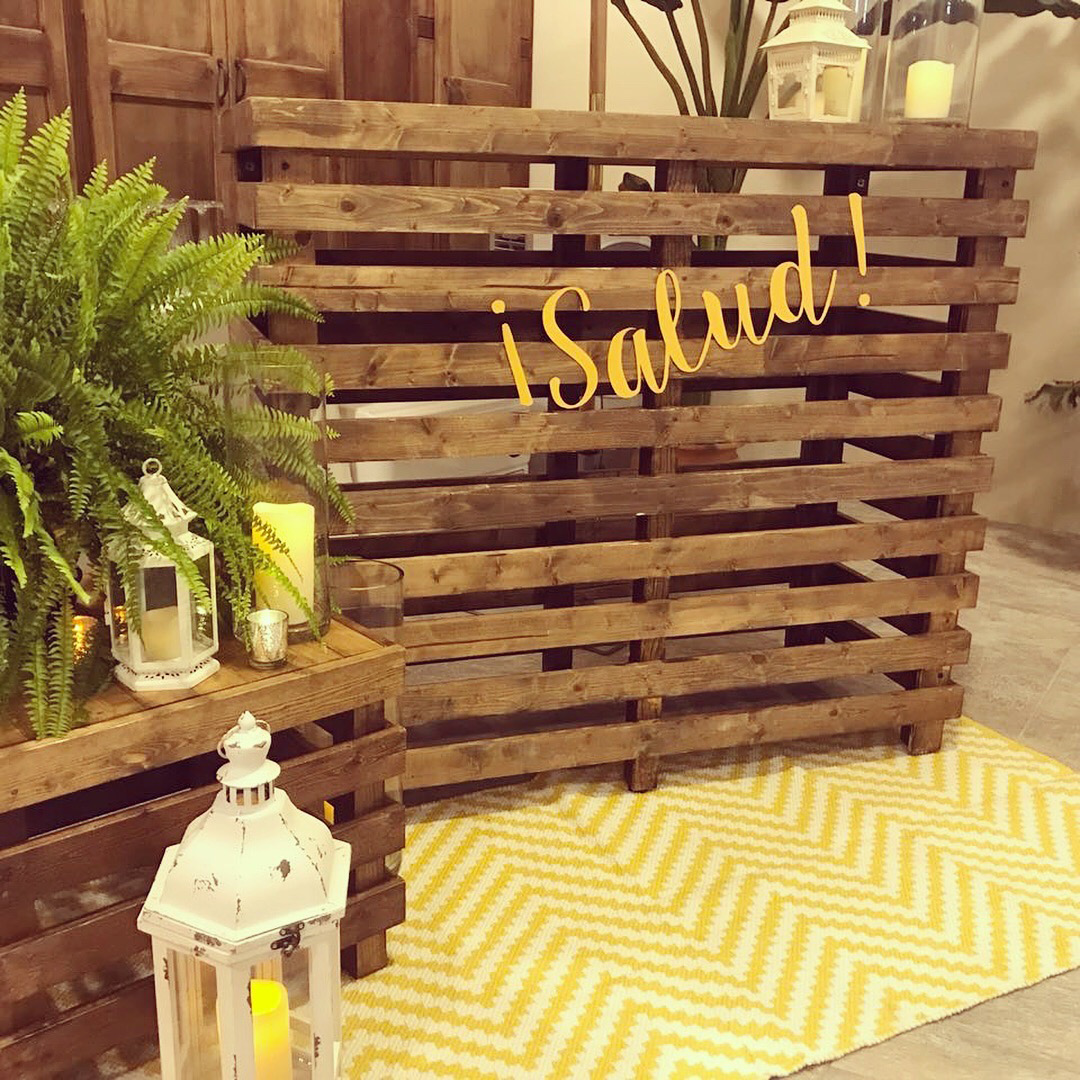 Pallet Bar Table by Trinidad Event Furniture features “¡Salud!” in yellow script, accented with candles, lanterns, a potted fern, and a yellow chevron-patterned rug. - Perched Event Rentals, Trinidad