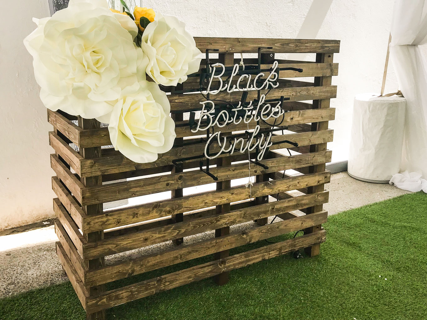 The Pallet Bar Table features a wooden pallet base with a neon "Black Bottles Only" sign and large white artificial flowers, set on artificial grass beside a white wall—ideal for Trinidad Wedding Hire or any chic event décor. - Perched Event Rentals, Trinidad