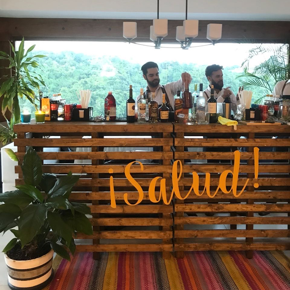Two bartenders prepare drinks behind a wooden bar with various liquor bottles. In the foreground, a potted plant and “¡Salud!” text are visible, showcasing the stylish Pallet Bar Table from Trinidad Event Hire. - Perched Event Rentals, Trinidad