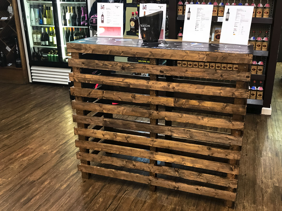 The Pallet Bar Table, ideal for Trinidad Event Hire, is displayed in a shop with menu cards placed on top, while shelves with bottles and a fridge can be seen in the background. - Perched Event Rentals, Trinidad