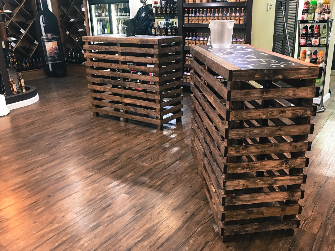 Two Pallet Bar Tables from Trinidad Event Rentals stand on a polished wooden floor in an off-licence, with shelves of bottles and a fridge in the background. - Perched Event Rentals, Trinidad