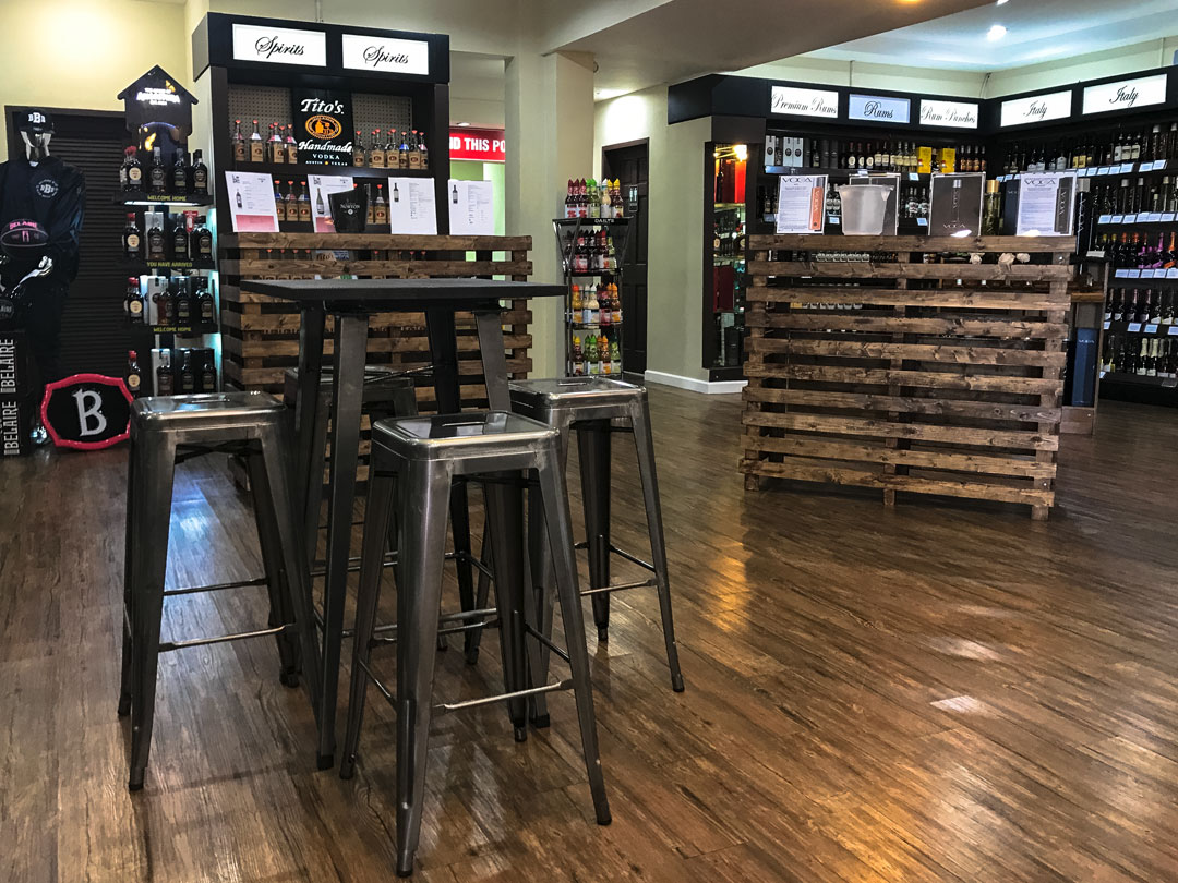 Interior of an off-licence with wooden floors, metal stools from Trinidad Event Furniture around a Pallet Bar Table, shelves of bottles, and wooden pallet counters. - Perched Event Rentals, Trinidad