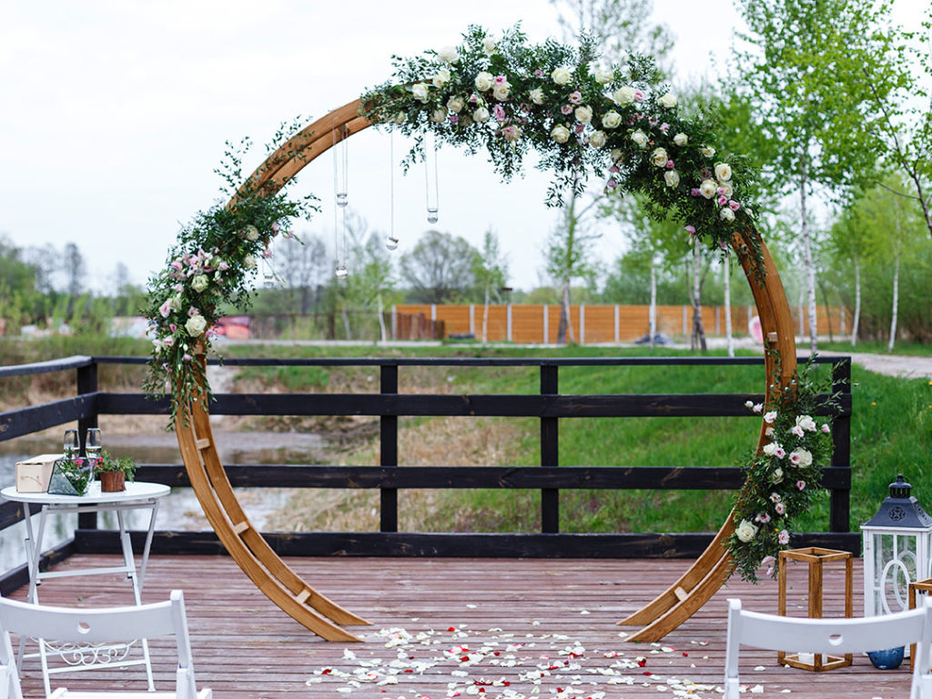 Oval Arch decorated with white and pink flowers and greenery on an outdoor wooden platform, with chairs and petals in front