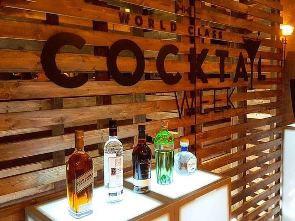 A Pallet Wall Backdrop with “World Class Cocktail Week” in large black letters, and five liquor bottles on a lit counter