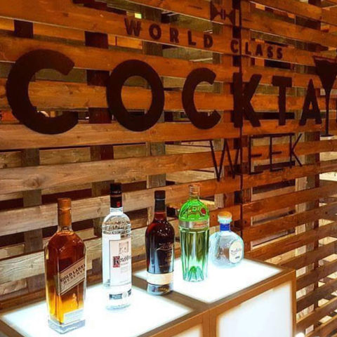 A Pallet Wall Backdrop with “World Class Cocktail Week” in large black letters, and five liquor bottles on a lit counter