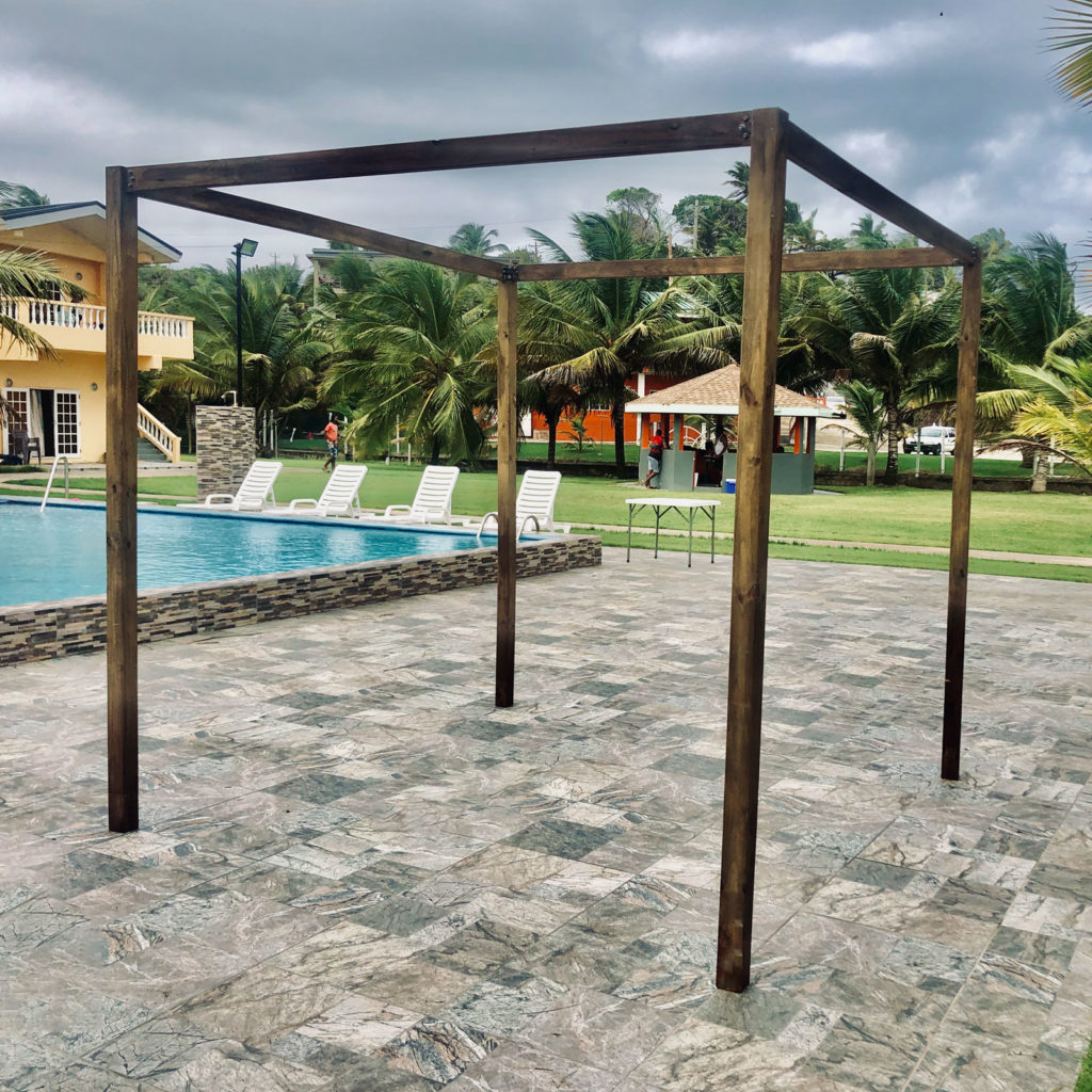 A Pergola Arch standing on a tiled poolside patio with loungers, a swimming pool and palm trees behind it under a cloudy sky
