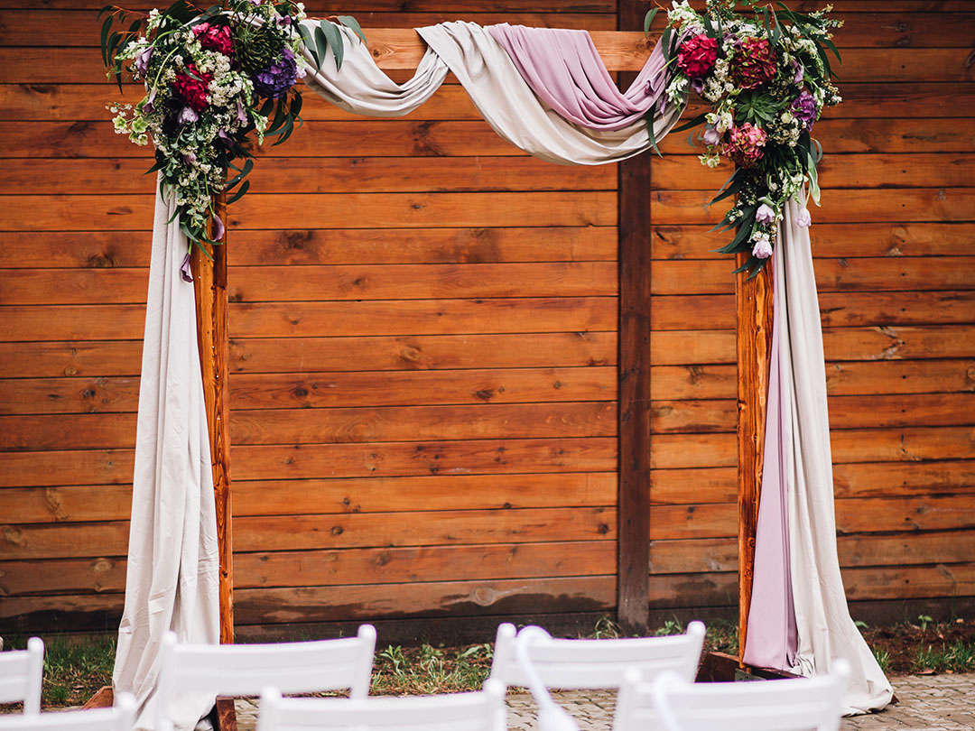 A Ceremony Arch draped with white and mauve fabric, adorned with red and purple flowers, stands before a wooden fence, with white chairs from Trinidad Event Hire arranged in front. - Perched Event Rentals, Trinidad