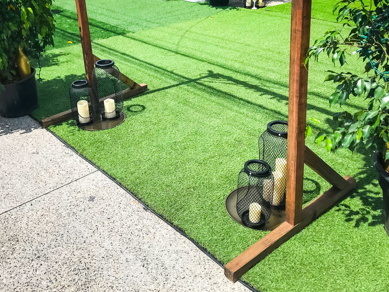 A stylish Ceremony Arch from Trinidad Event Rentals frames two black metal lanterns with white candles on artificial grass by wooden posts and a concrete path, adding elegance to the setting. - Perched Event Rentals, Trinidad