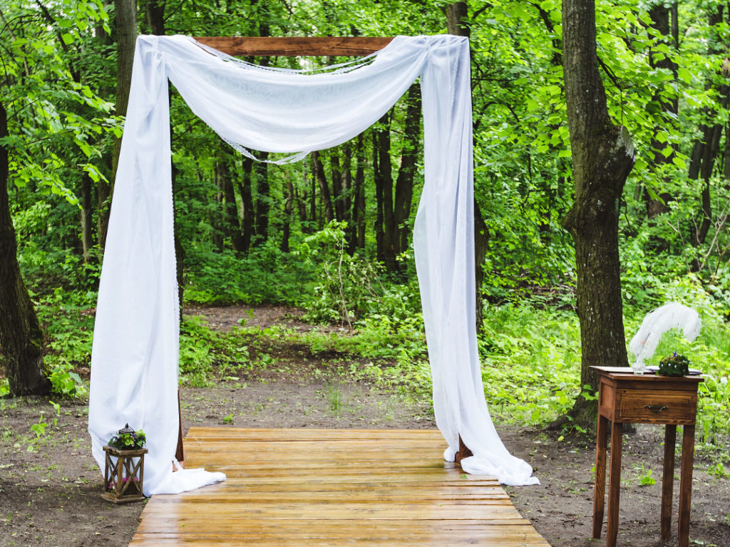 The Ceremony Arch draped with white fabric on a wooden walkway in a green forest, with a small table and a lantern beside it