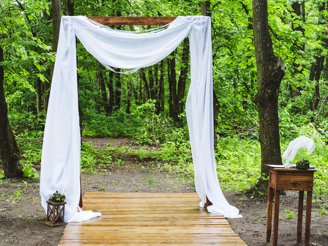The Ceremony Arch draped with white fabric on a wooden walkway in a green forest, with a small table and a lantern beside it