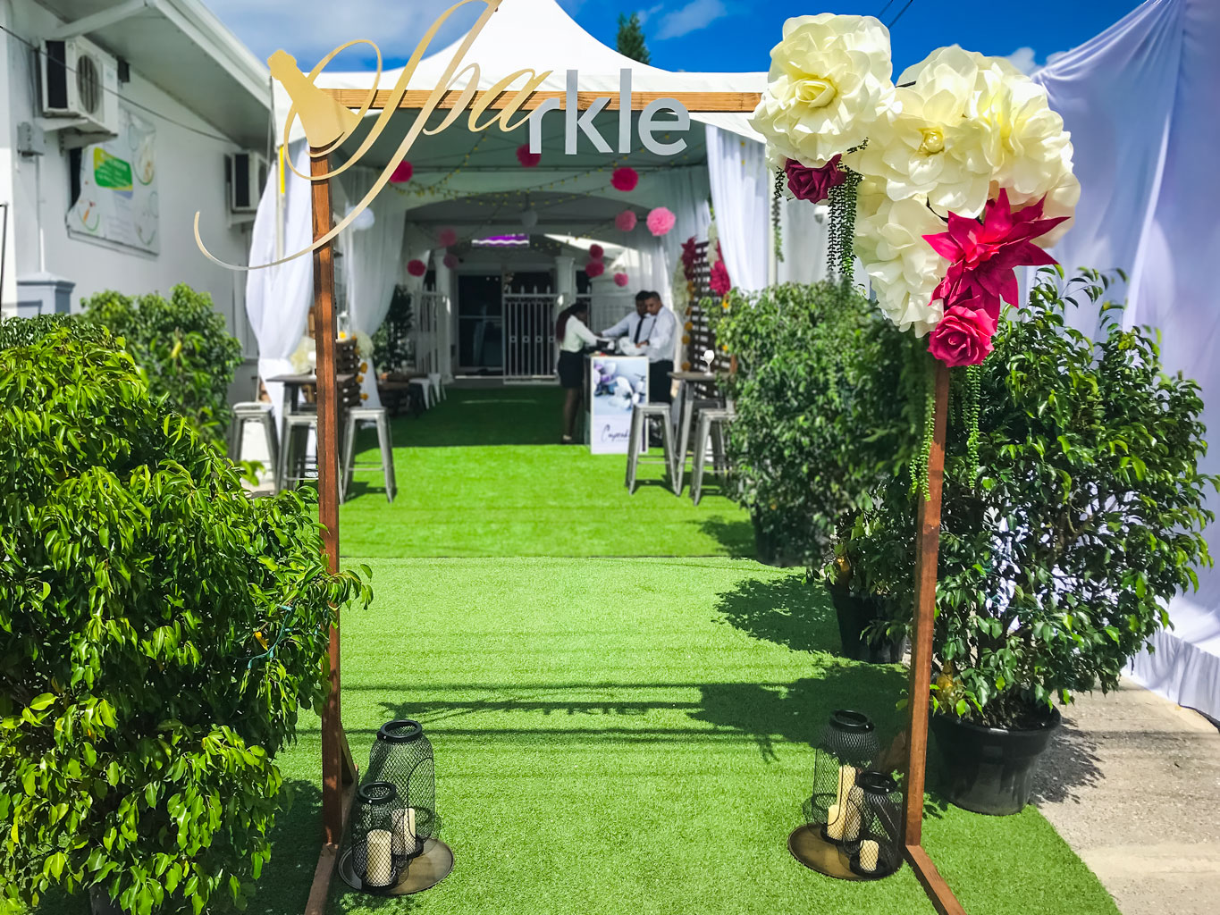 Entrance to an outdoor event features artificial grass, decorative lanterns, and a Ceremony Arch leading to a tented seating area with Trinidad Event Furniture. Two people stand inside near a sign. - Perched Event Rentals, Trinidad