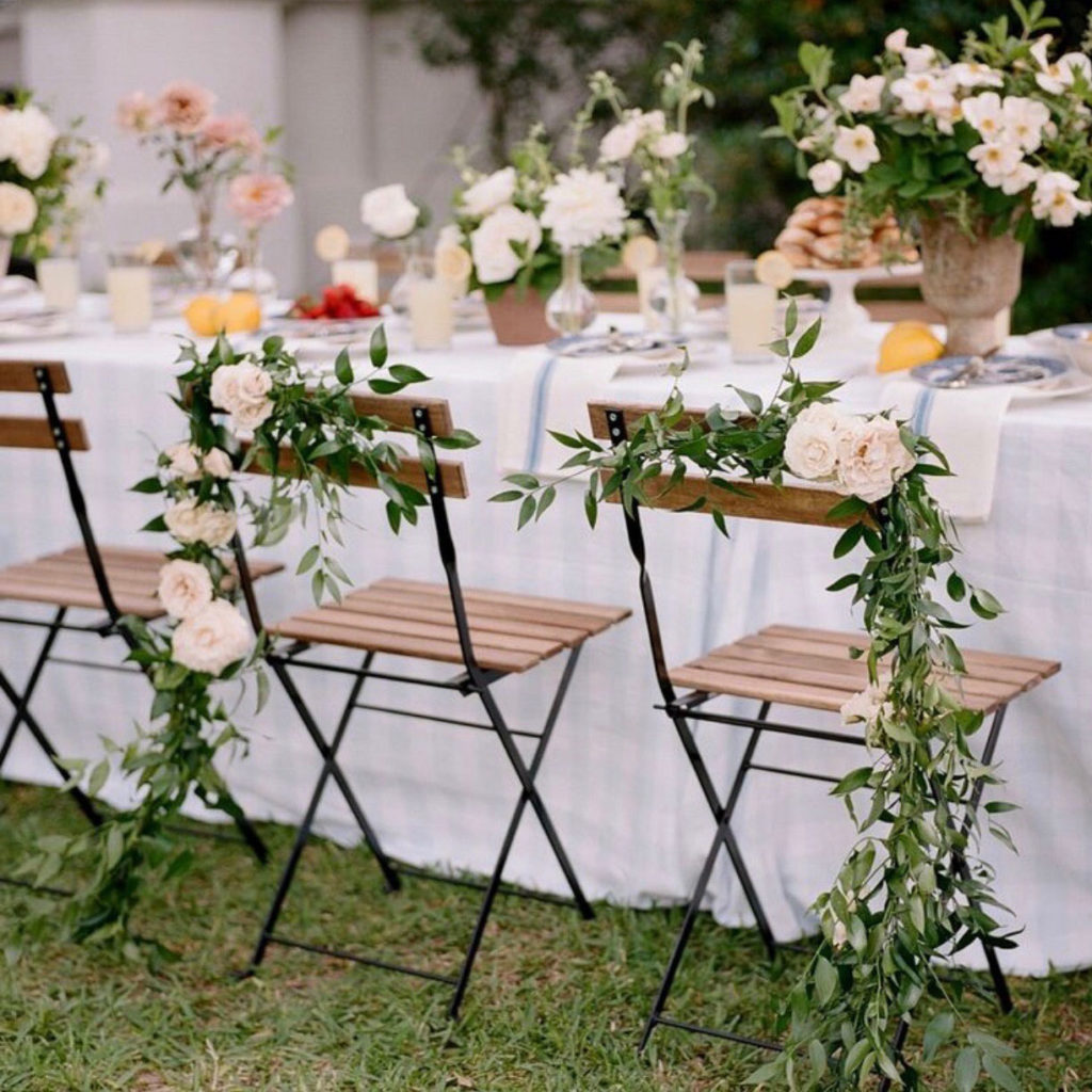 An outdoor table with French Bistro Chairs draped in greenery and white flowers, floral centrepieces, plates and glasses