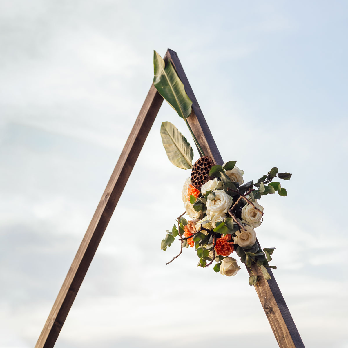 The A-Frame Arch from Trinidad Wedding Hire, adorned with white and orange flowers and green leaves, stands beautifully against a cloudy sky. - Perched Event Rentals, Trinidad