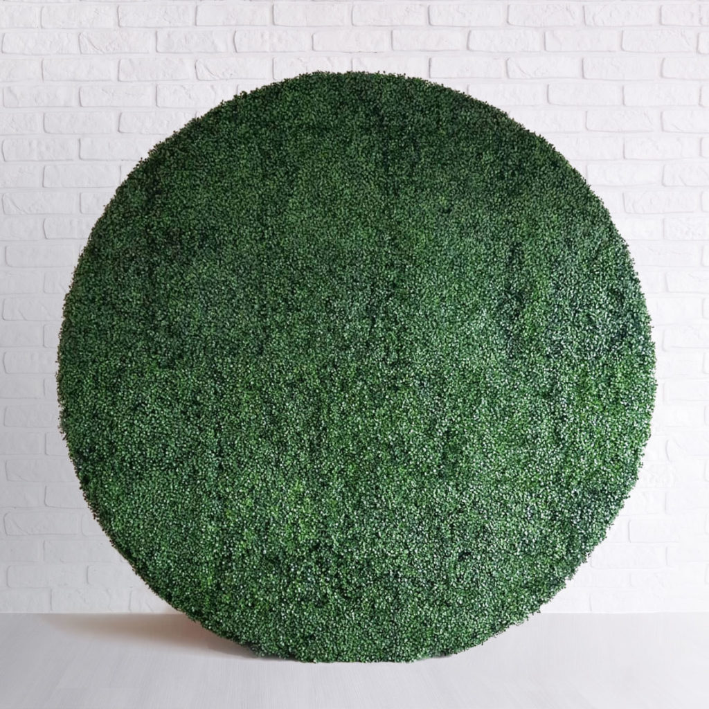 Circular Boxwood Backdrop, a large round artificial green hedge, against a white brick wall on a light floor