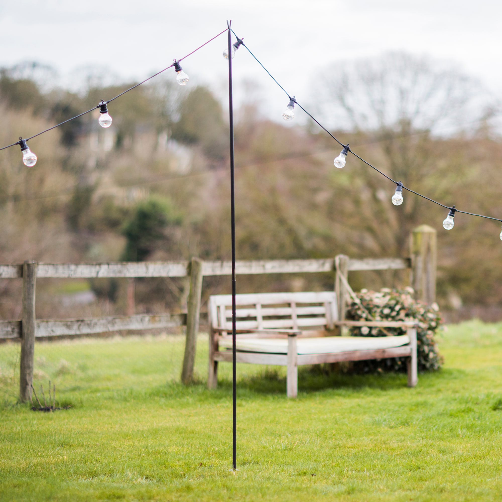 A metal pole from Trinidad Event Rentals holds up a string of outdoor lights on a grassy lawn, with a wooden bench and fence in the background. - Perched Event Rentals, Trinidad